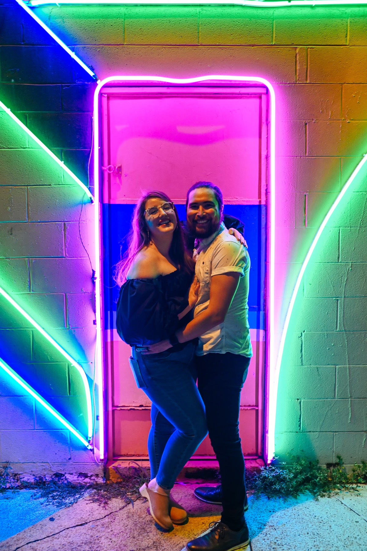 Nick and Colleen embracing and smiling at the camera in front of a neon art installation