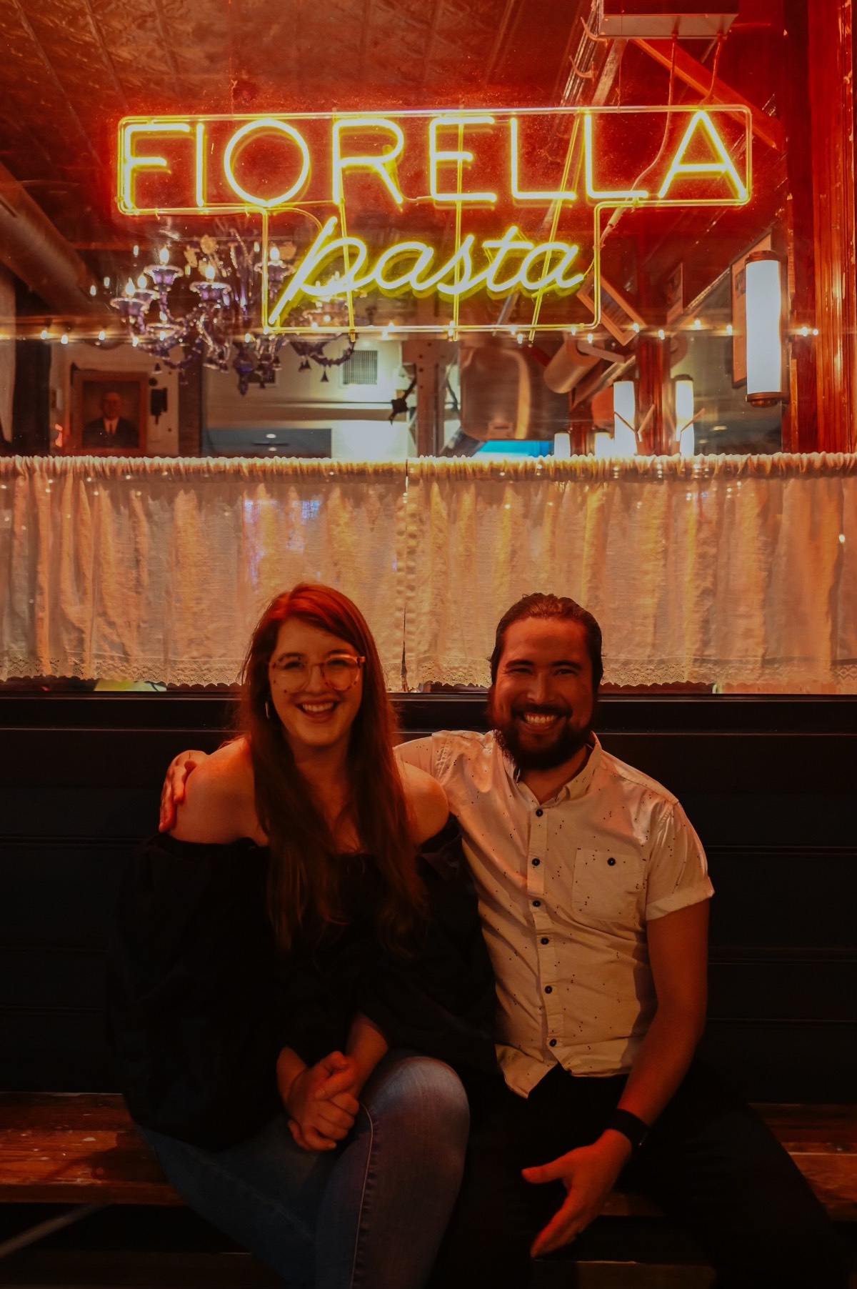 Nick and Colleen smiling at the camera and sitting on a bench in front of a lit sign that reads “Fiorella Pasta”